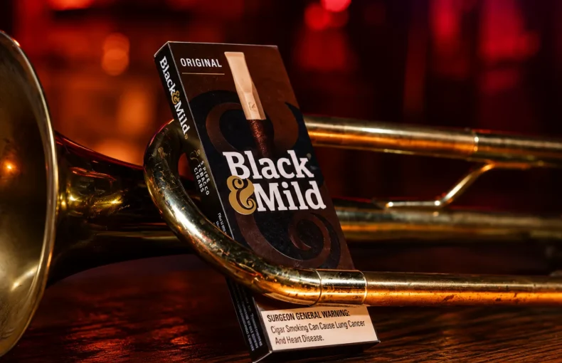 Close-up of a Black & Mild Original cigar box resting on a brass trumpet in a moody red-lit setting, warning label visible on the box's edge.