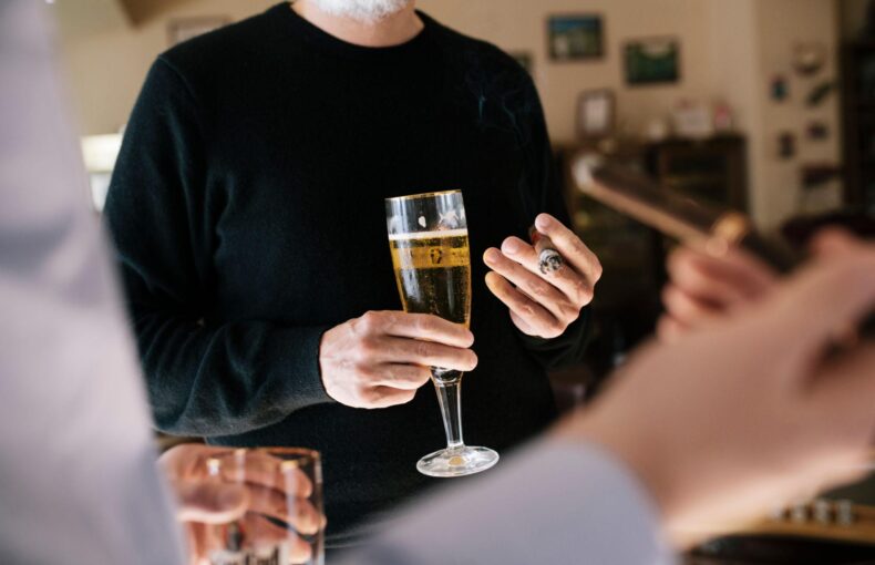 Person in a black shirt holding a tall champagne flute and a lit cigar at a social gathering indoors.