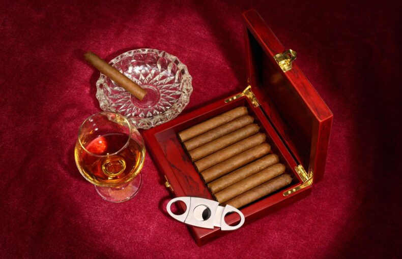 Open red wooden cigar box with cigars, a glass of whiskey, and a crystal ashtray on a burgundy velvet surface.
