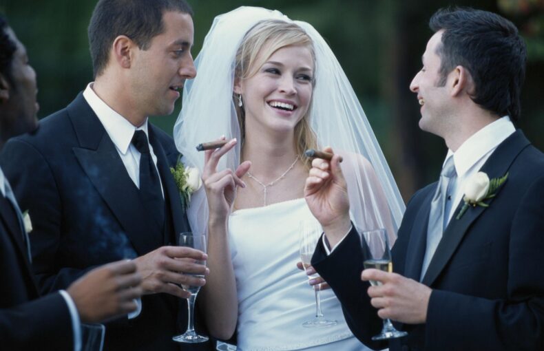 Bride in a white wedding gown and veil laughing, flanked by two men in tuxedos, all toasting with champagne outdoors.