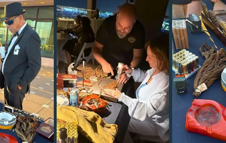 Two men in business attire at an outdoor reception examine a cigar display on a table, cups in hand. If context matters, they’re inspecting a row of cigars on a tray beside other beverages.